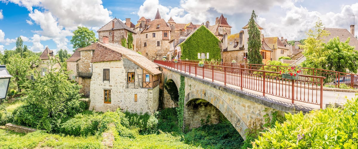 A stone bridge with a red railing, surrounded by lush greenery and historic stone buildings.