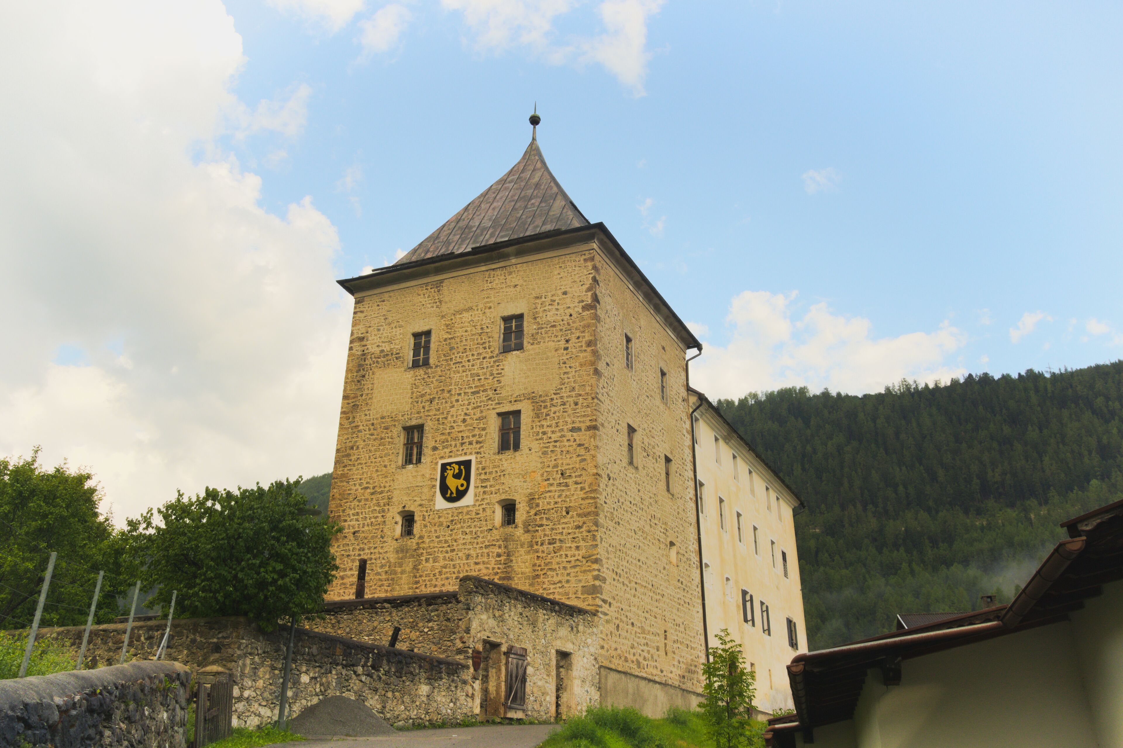 A stone tower with a pointed roof, a crest, and a forested hill in the background.