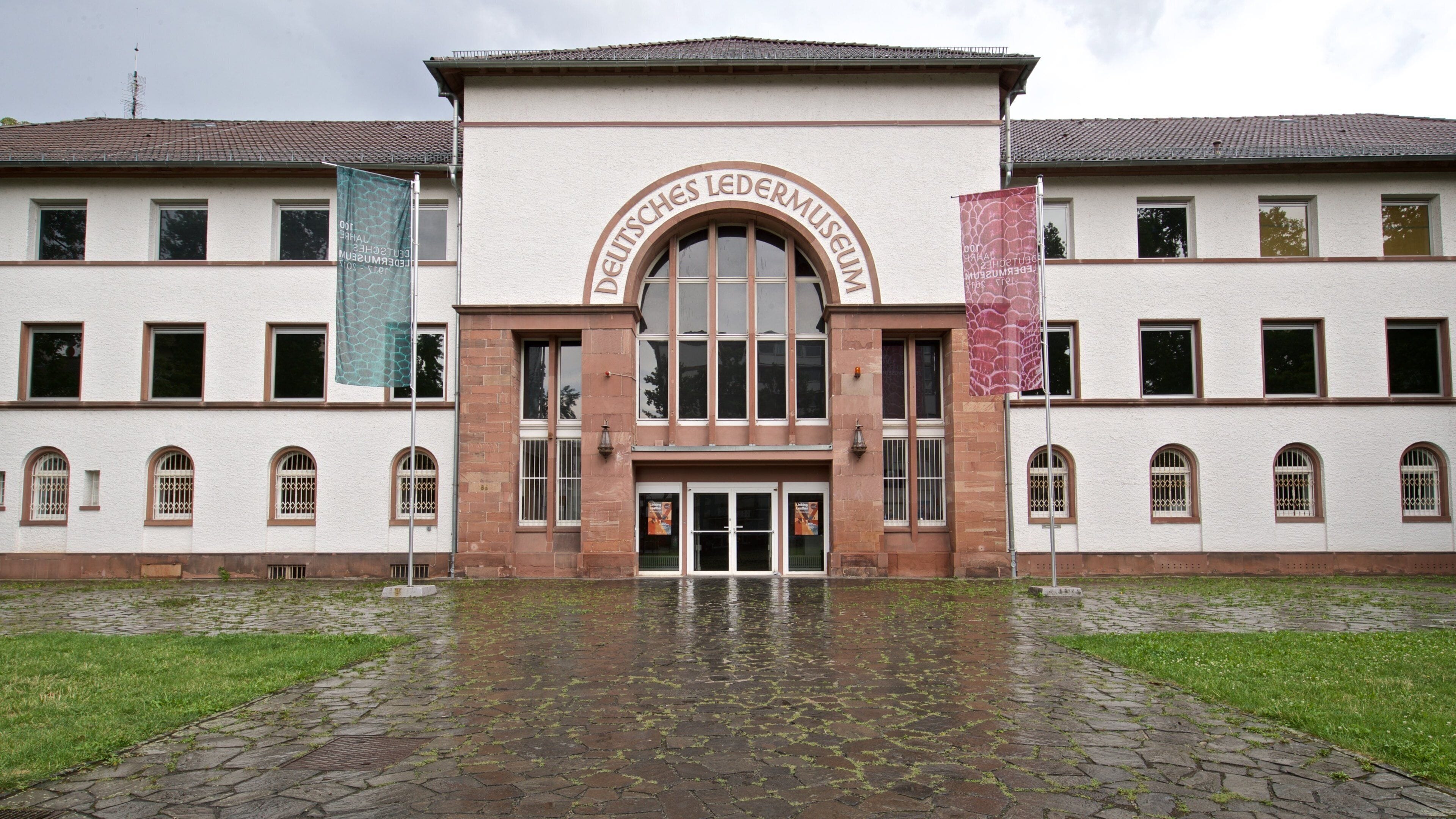 A entrada do Deutsches Ledermuseum, com telhado de azulejos, janelas em arco e duas bandeiras de cada lado.