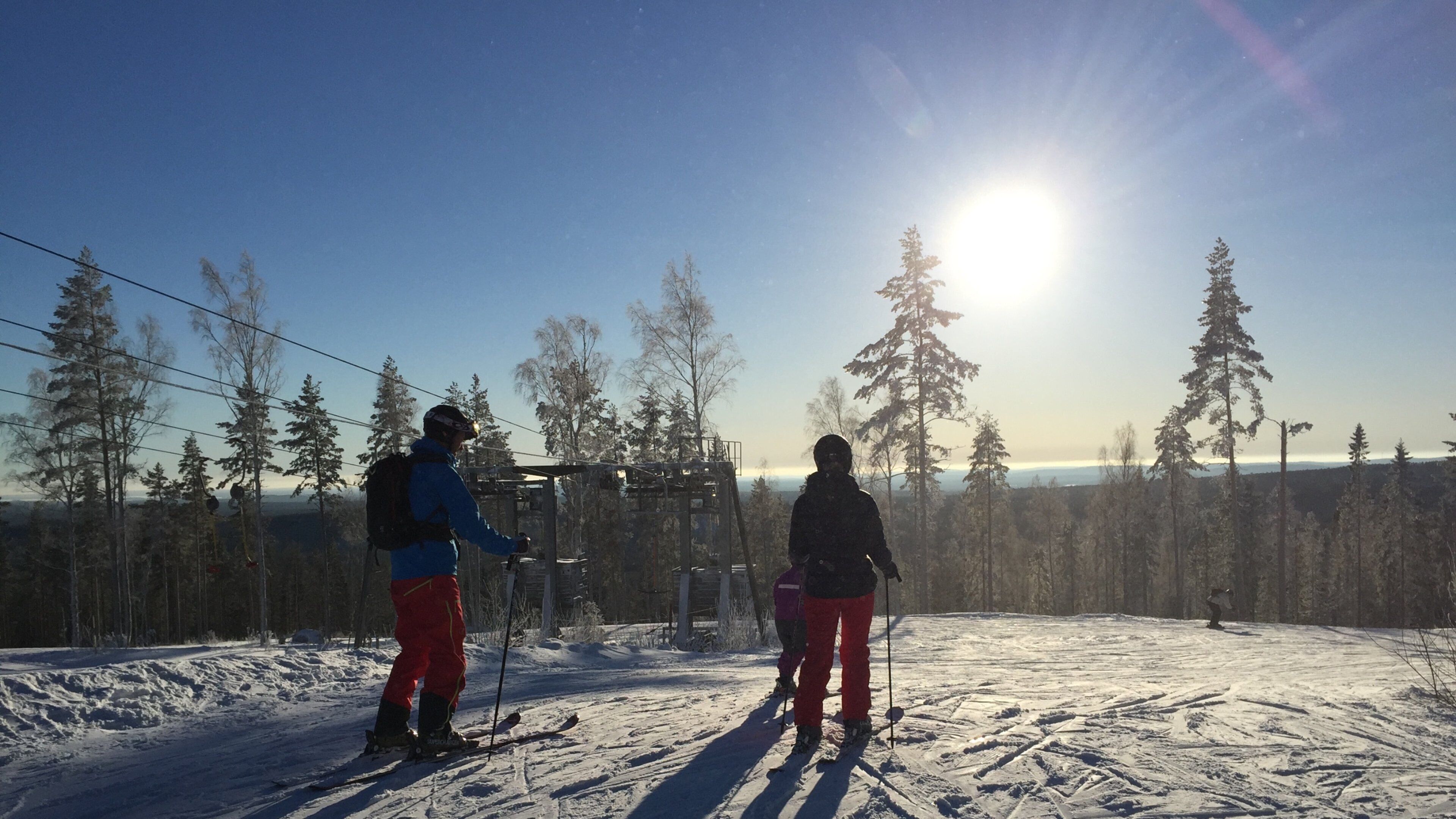 Two skiers on a snowy slope with a clear sky and a sun in the background.