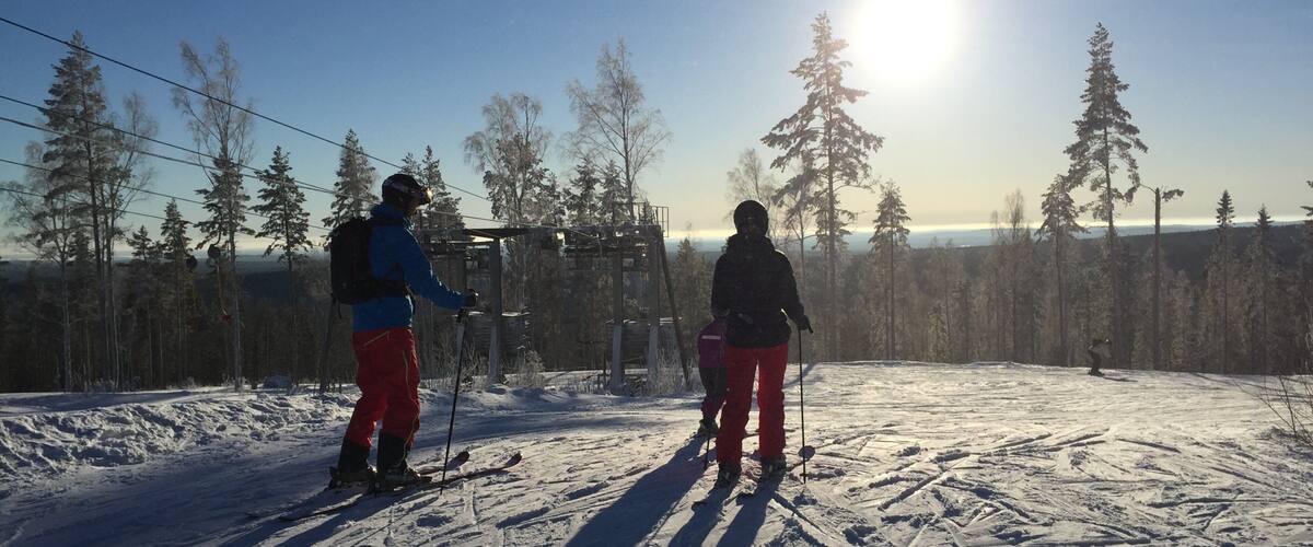 Two skiers on a snowy slope with a clear sky and a sun in the background.