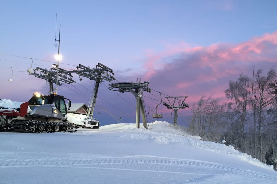 Un telesilla cubierto de nieve con huellas en la nieve, una cabaña de madera y un bosque de árboles sin hojas bajo un cielo rosado y morado.