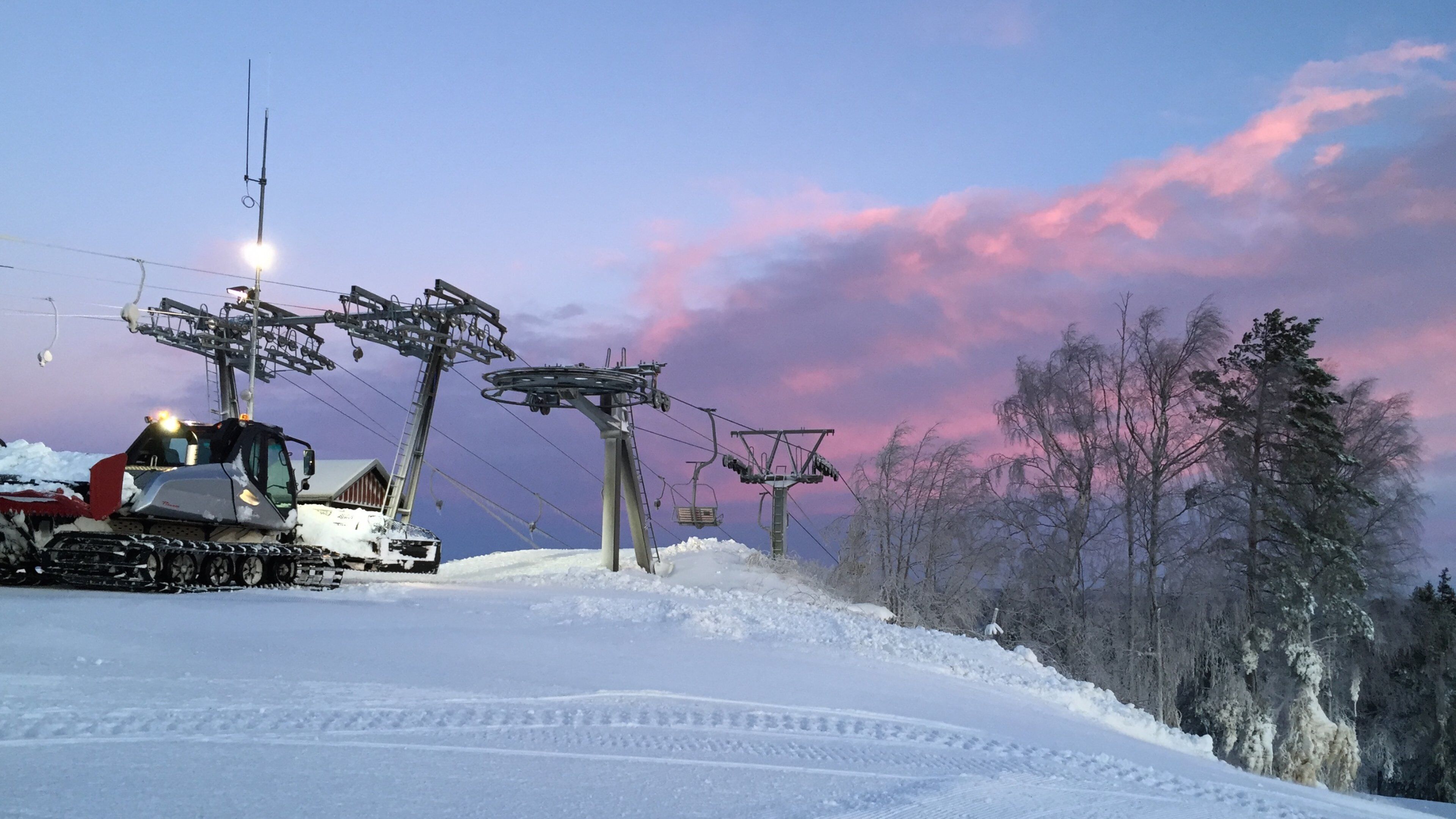 A snow-covered ski lift with tracks in the snow, a wooden cabin, and a forest of leafless trees under a pink and purple sky.