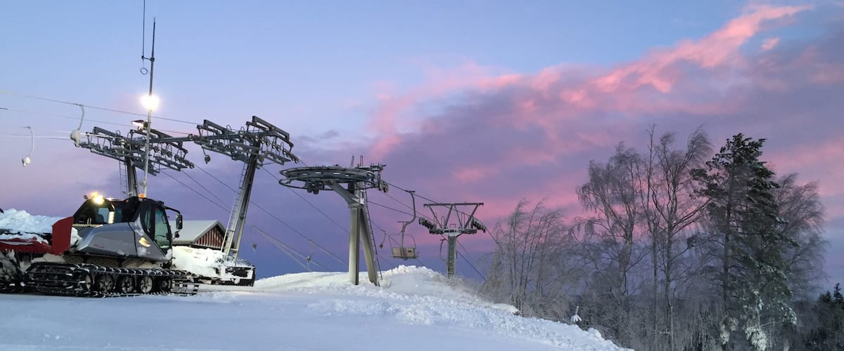 A snow-covered ski lift with tracks in the snow, a wooden cabin, and a forest of leafless trees under a pink and purple sky.