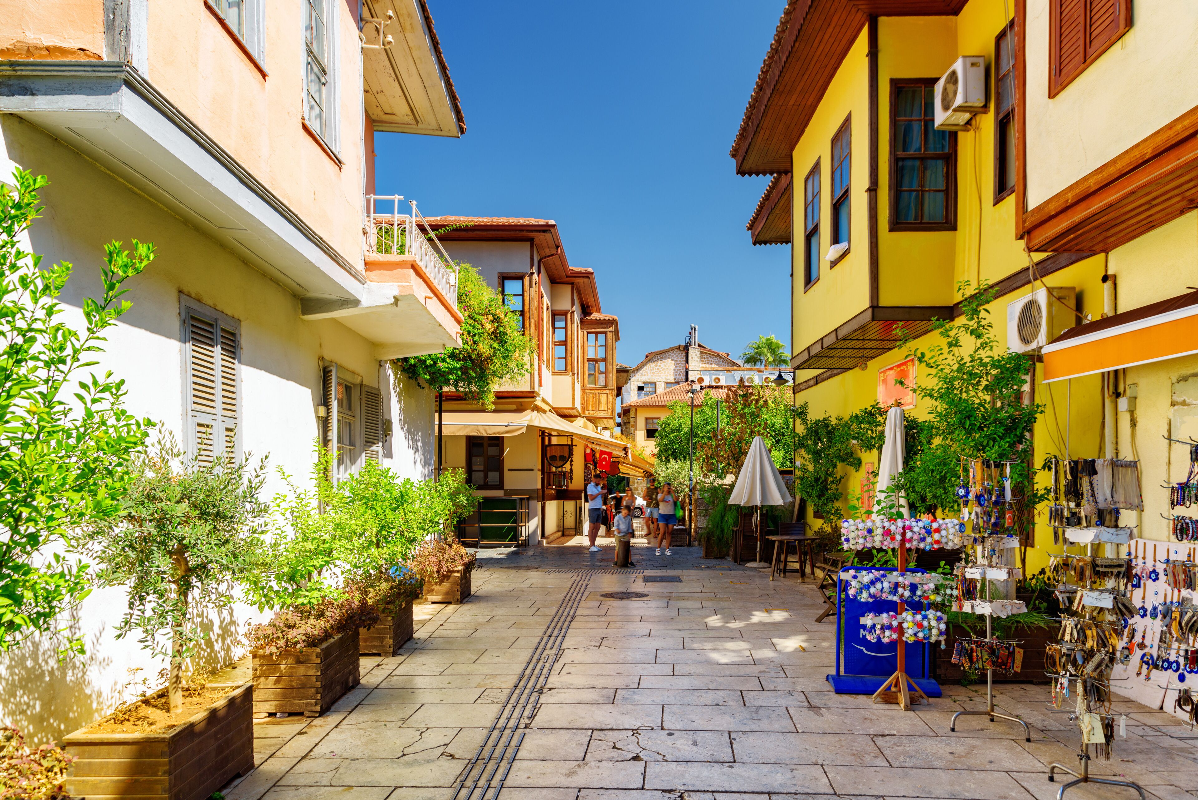 A narrow street lined with colorful buildings, potted plants, and outdoor seating areas.