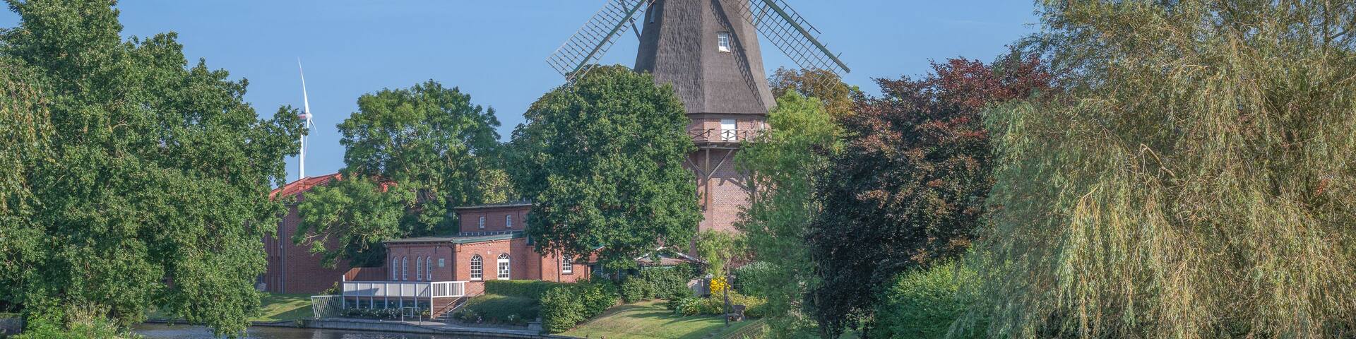 A traditional windmill by a river, surrounded by lush greenery and a brick building.