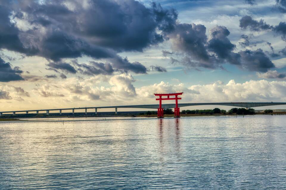 A red torii gate in the water with a bridge in the background.