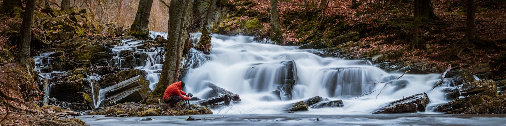 A waterfall in a forest with a person in a red jacket taking a photo.