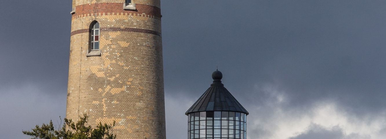 Un alto faro rojo y verde con base de ladrillo, rodeado de árboles y un cielo nublado.