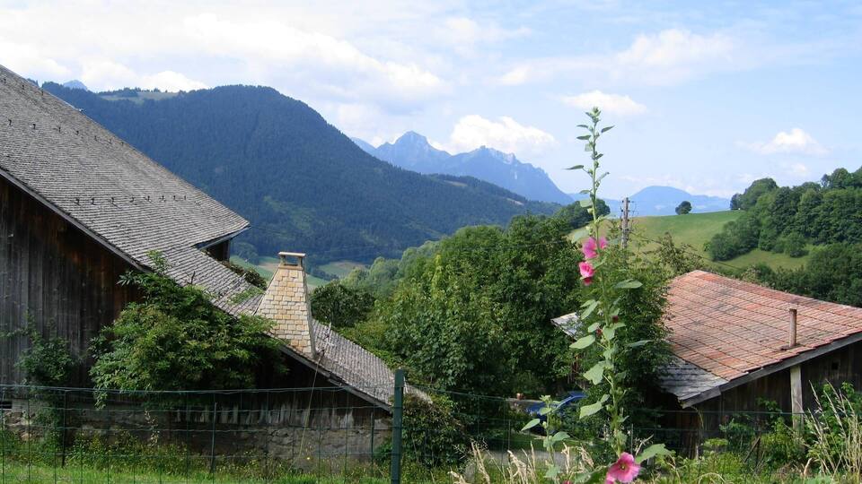 A rural landscape with a wooden house, a stone chimney, and a mountain range in the background.