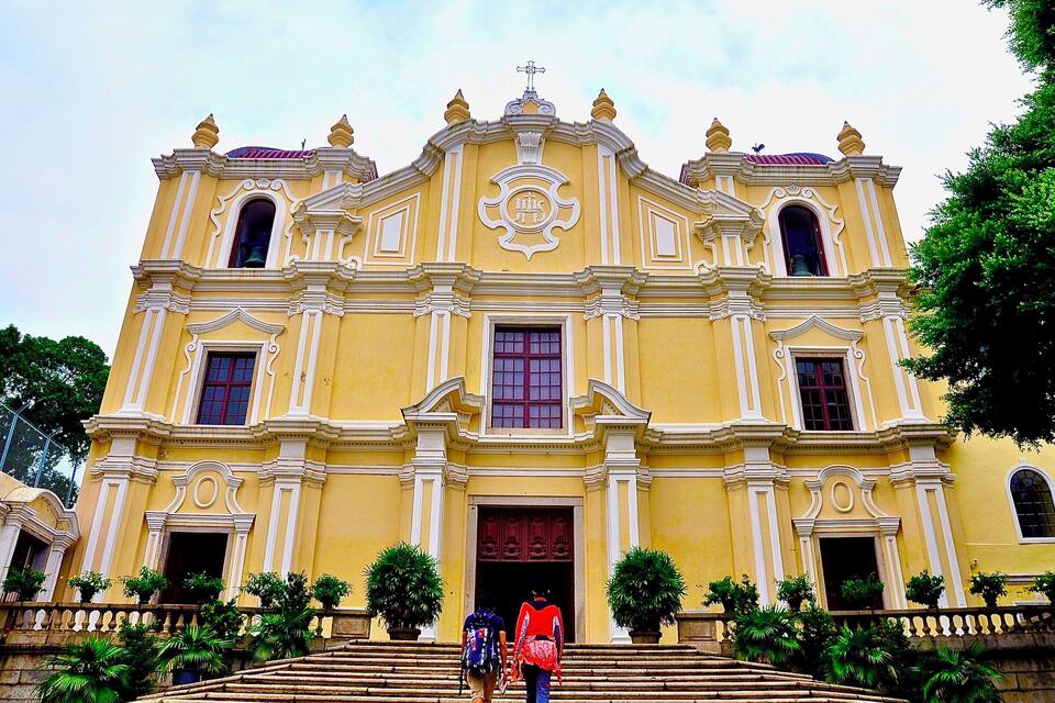A yellow colonial-style church with a bell tower, steps leading up to the entrance, and potted plants in front.