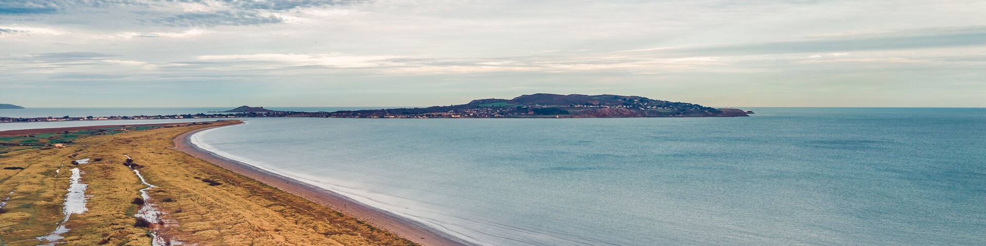 A coastal landscape with a sandy beach, rocky intertidal zone, and a distant hill.
