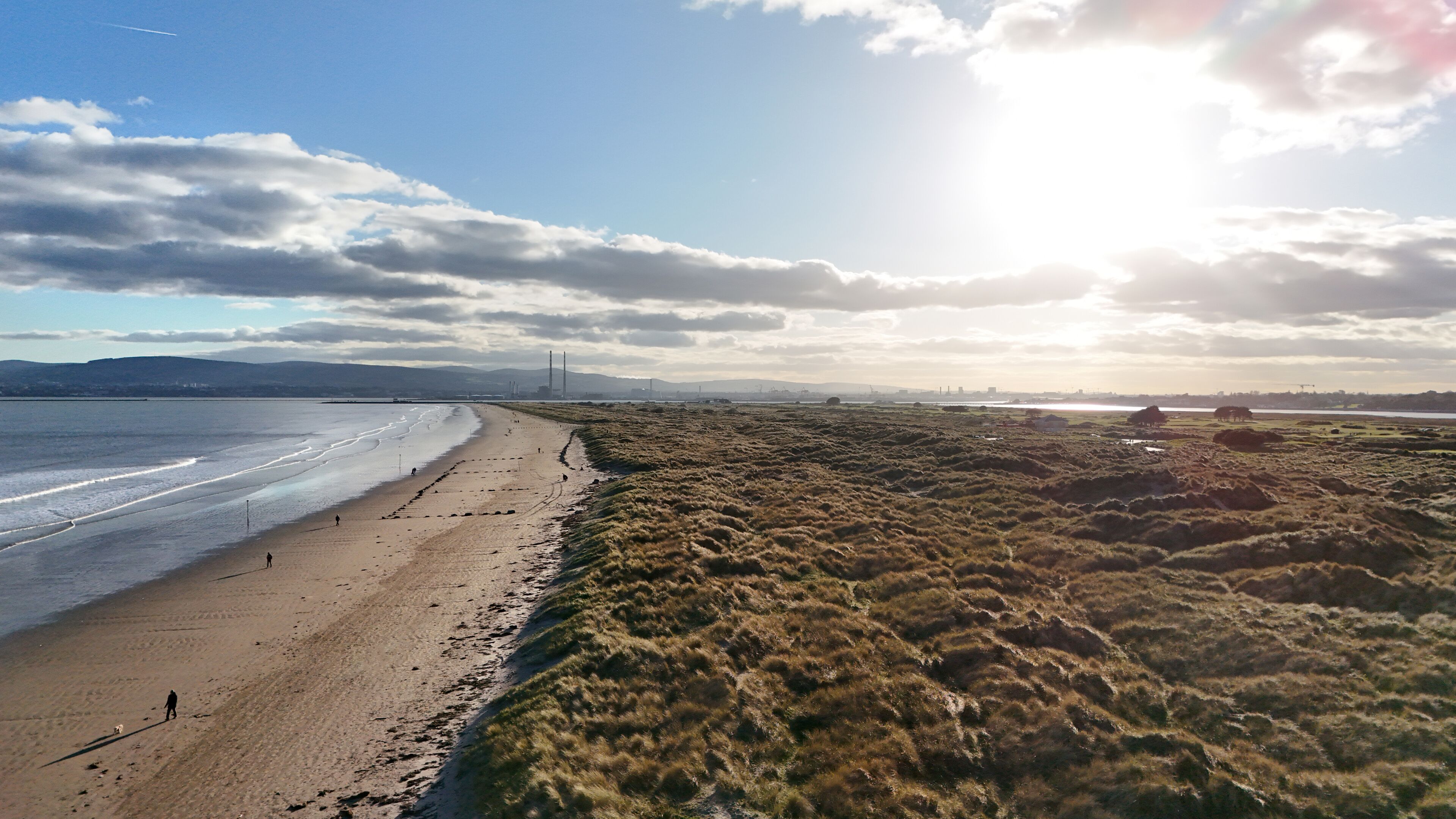 A beach with a grassy area beside it, a distant bridge, and a cloudy sky.