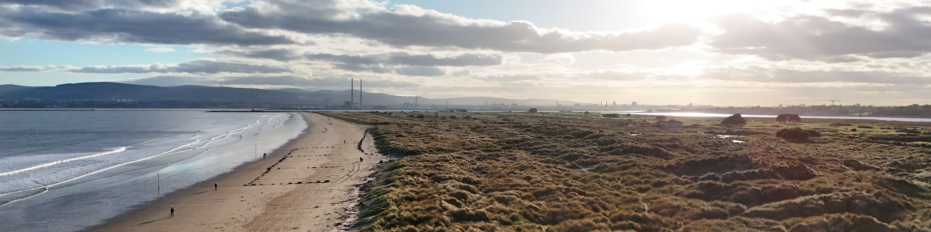 A beach with a grassy area beside it, a distant bridge, and a cloudy sky.