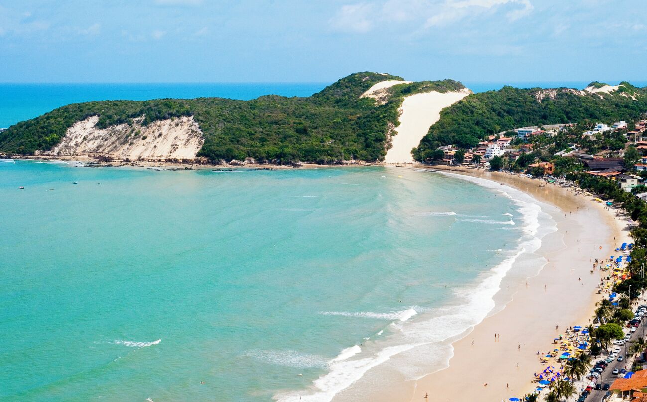 Uma praia costeira com águas turquesa cristalinas, areal arenoso e uma área densamente povoada com edifícios e palmeiras.