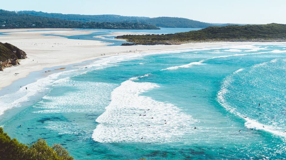 A coastal landscape with clear turquoise waters, sandy beaches, and a forested hill in the background.