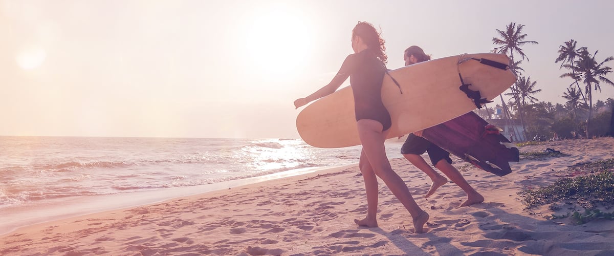 A beach with palm trees and people carrying surfboards.