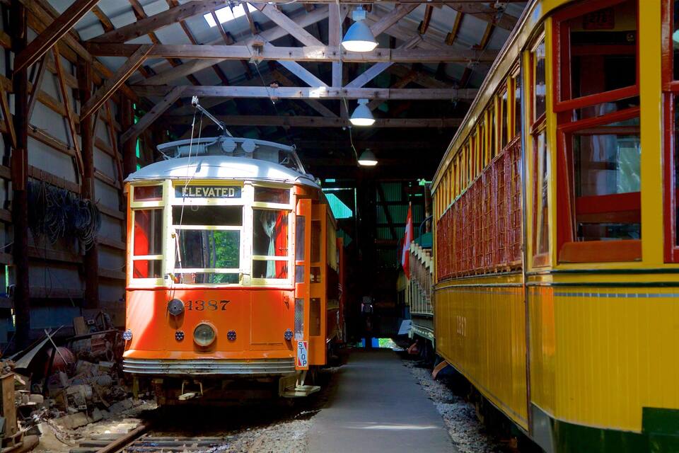 Un tramway ancien, aux couleurs orange et jaune, se trouve à l’intérieur d’une structure couverte, avec un plafond en bois et des poutres métalliques.