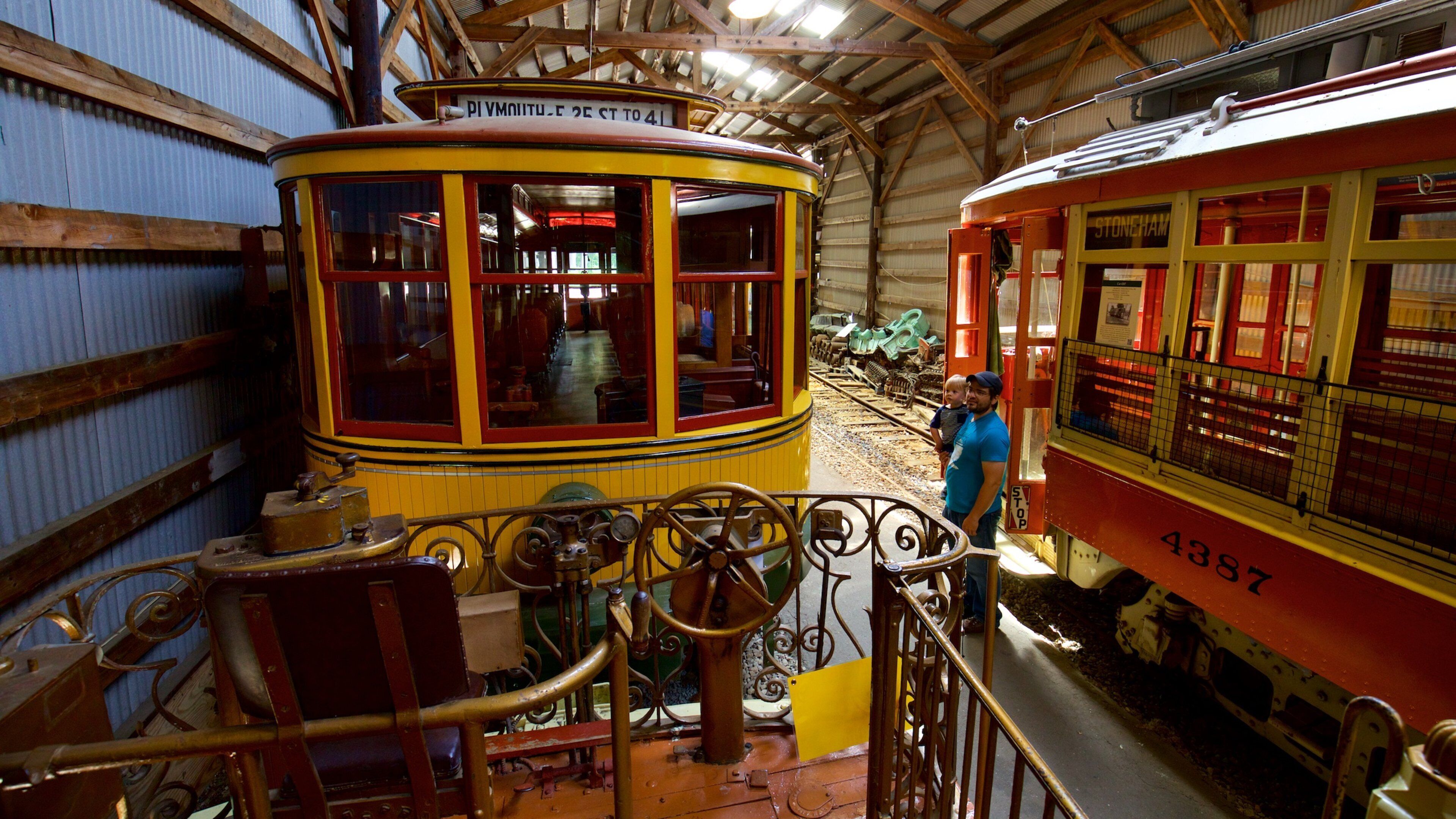 A yellow and red vintage tram with a sign reading "Stoneham 2E to 4I" inside a building with wooden beams and corrugated metal walls.