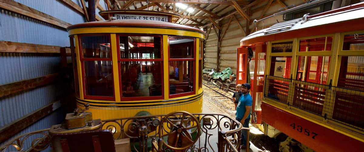 A yellow and red vintage tram with a sign reading "Stoneham 2E to 4I" inside a building with wooden beams and corrugated metal walls.