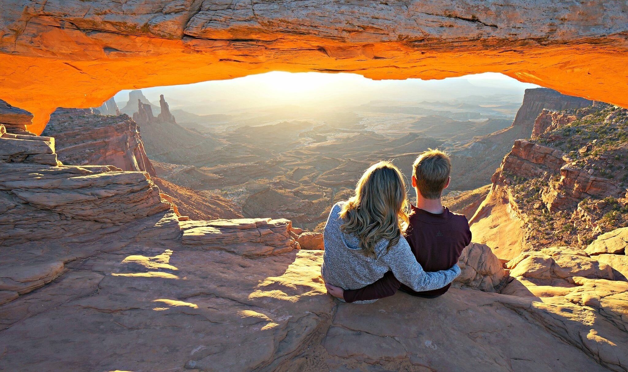 Una pareja sentada sobre una roca con vista a un cañón que tiene una formación arqueada.