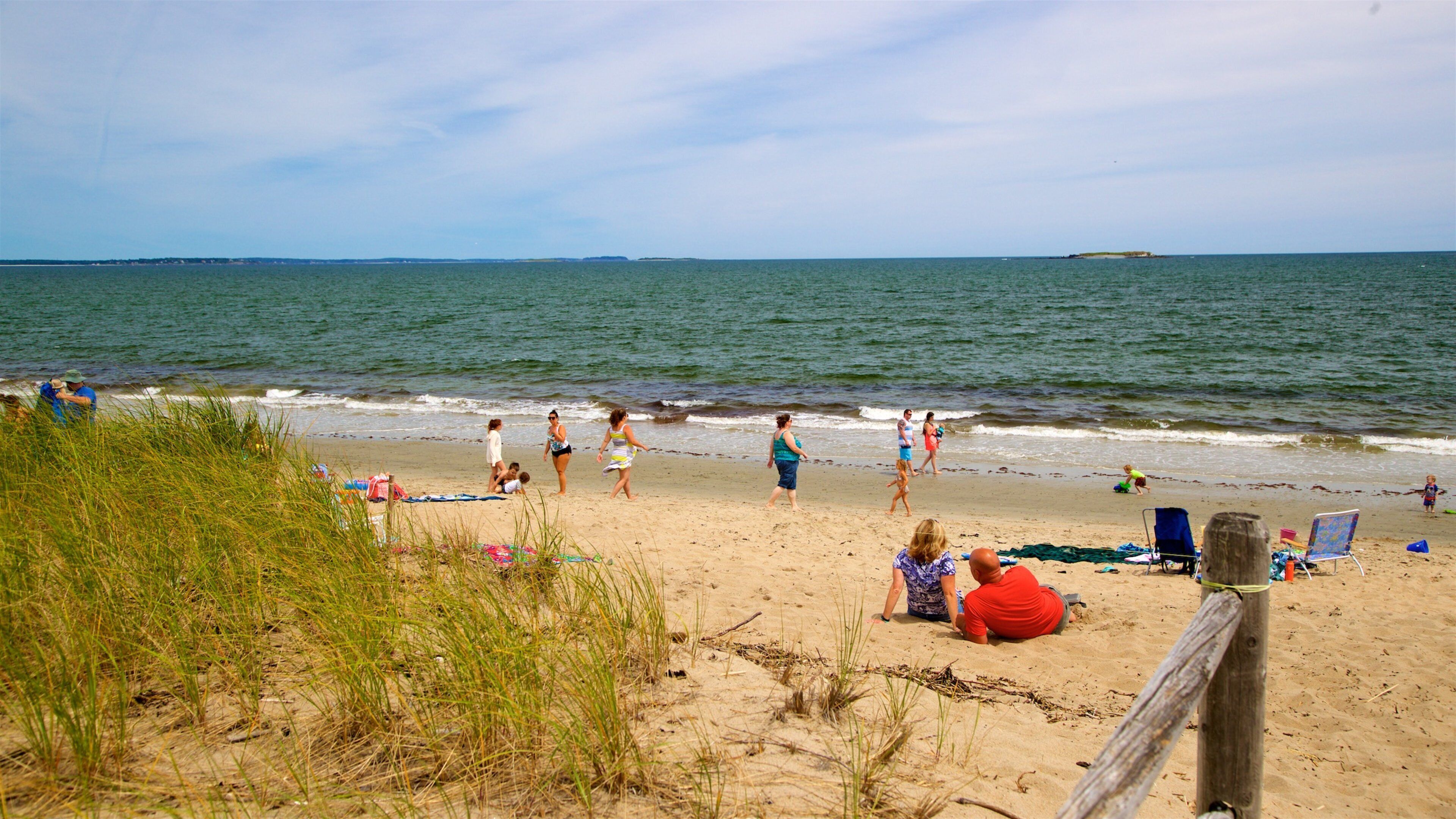 A beach with people walking and sitting on the sand, a wooden fence, and the ocean in the background.