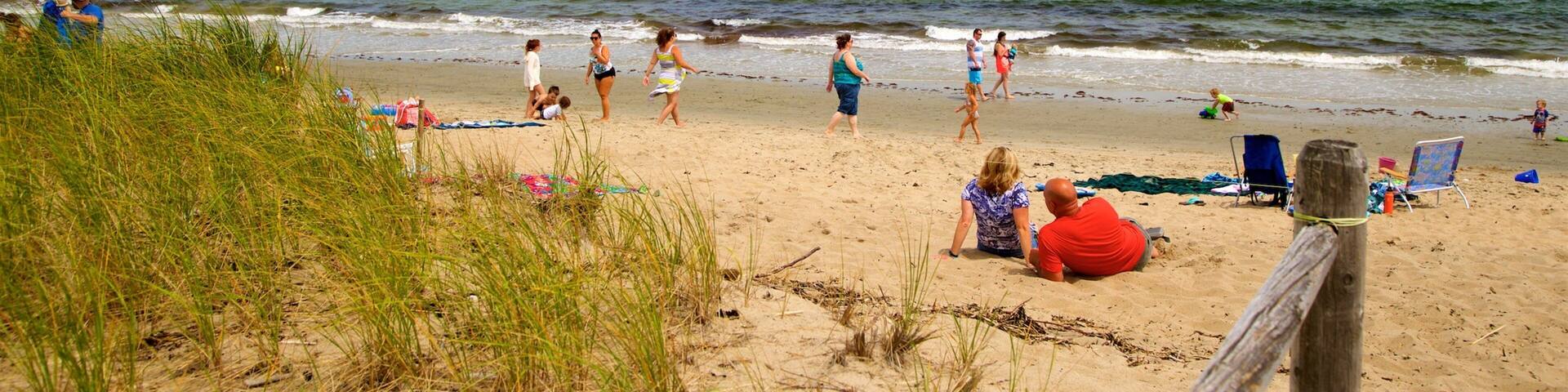 Una playa con gente caminando y sentada sobre la arena, una valla de madera y el océano al fondo.