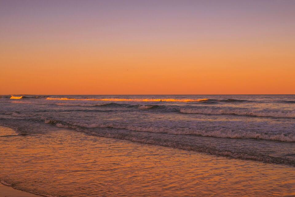 A beach at sunset with waves and two people carrying surfboards.