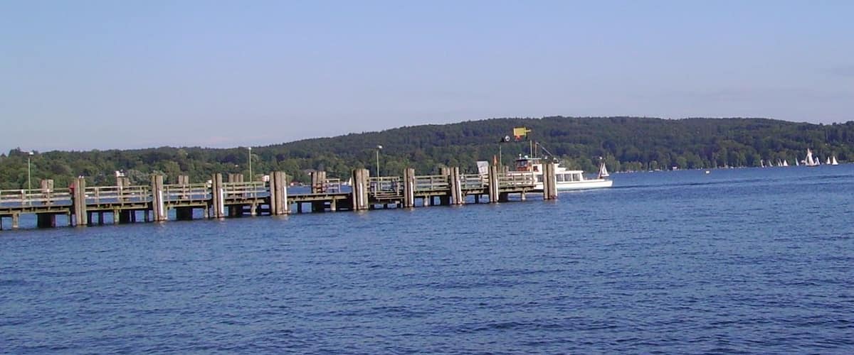 A wooden pier extending into a calm body of water with a white boat docked at the end. In the background, there are hills covered with trees and several sailboats on the water.