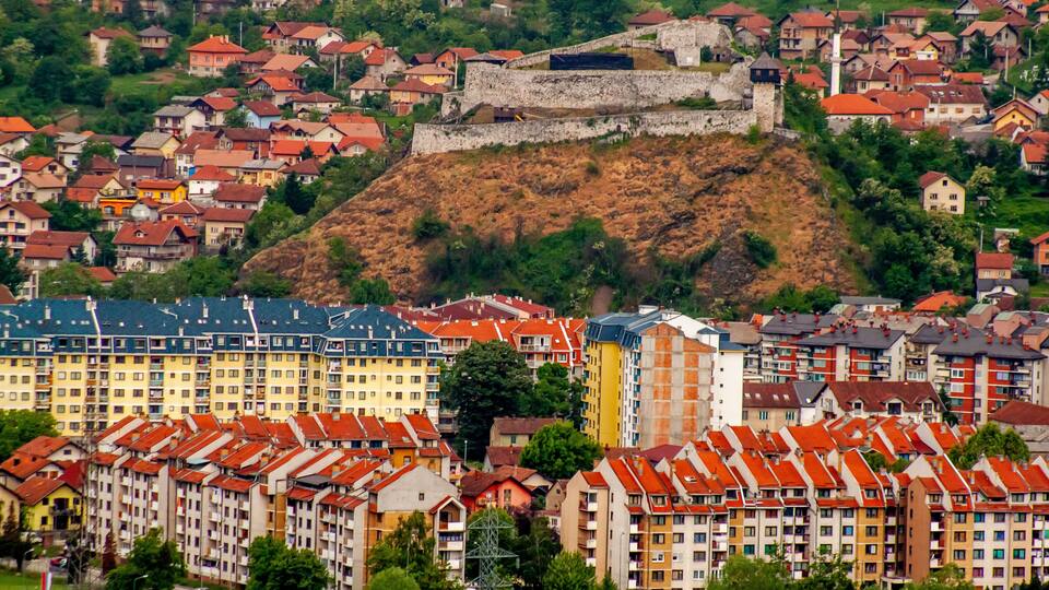 A town with colorful buildings, a prominent hill with a fort, and a river in the foreground.