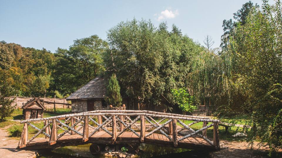 A wooden bridge over a calm river, surrounded by lush greenery and a small stone building.