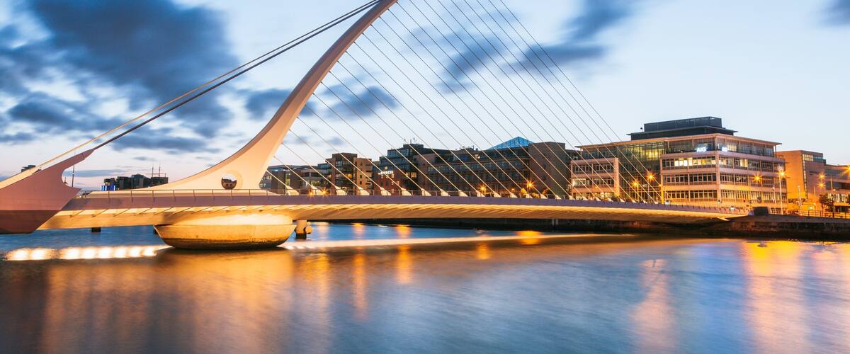 A modern bridge with a unique cable design, a body of water in the foreground, and a cityscape with buildings in the background.