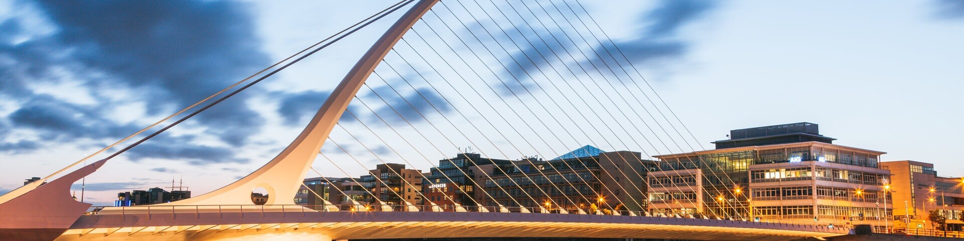 A modern bridge with a unique cable design, a body of water in the foreground, and a cityscape with buildings in the background.