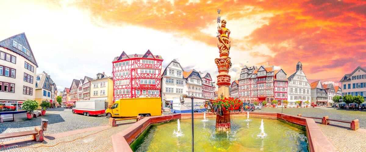 A town square with a fountain, traditional half-timbered buildings, and a truck.
