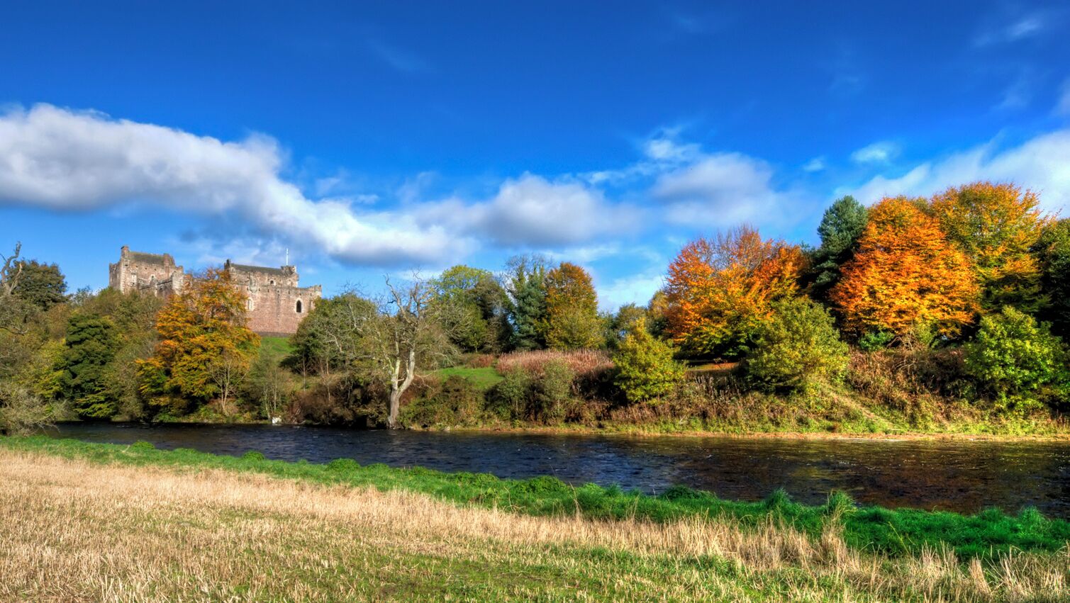 A river with a castle in the background, surrounded by autumn-colored trees and a clear blue sky.