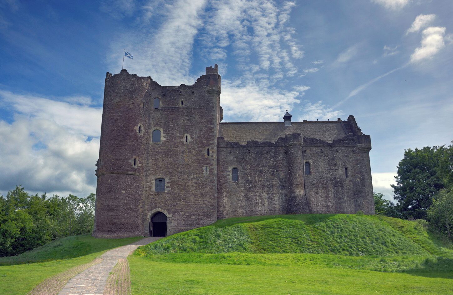 A historic stone castle with a flag, surrounded by lush green hills and a clear sky.