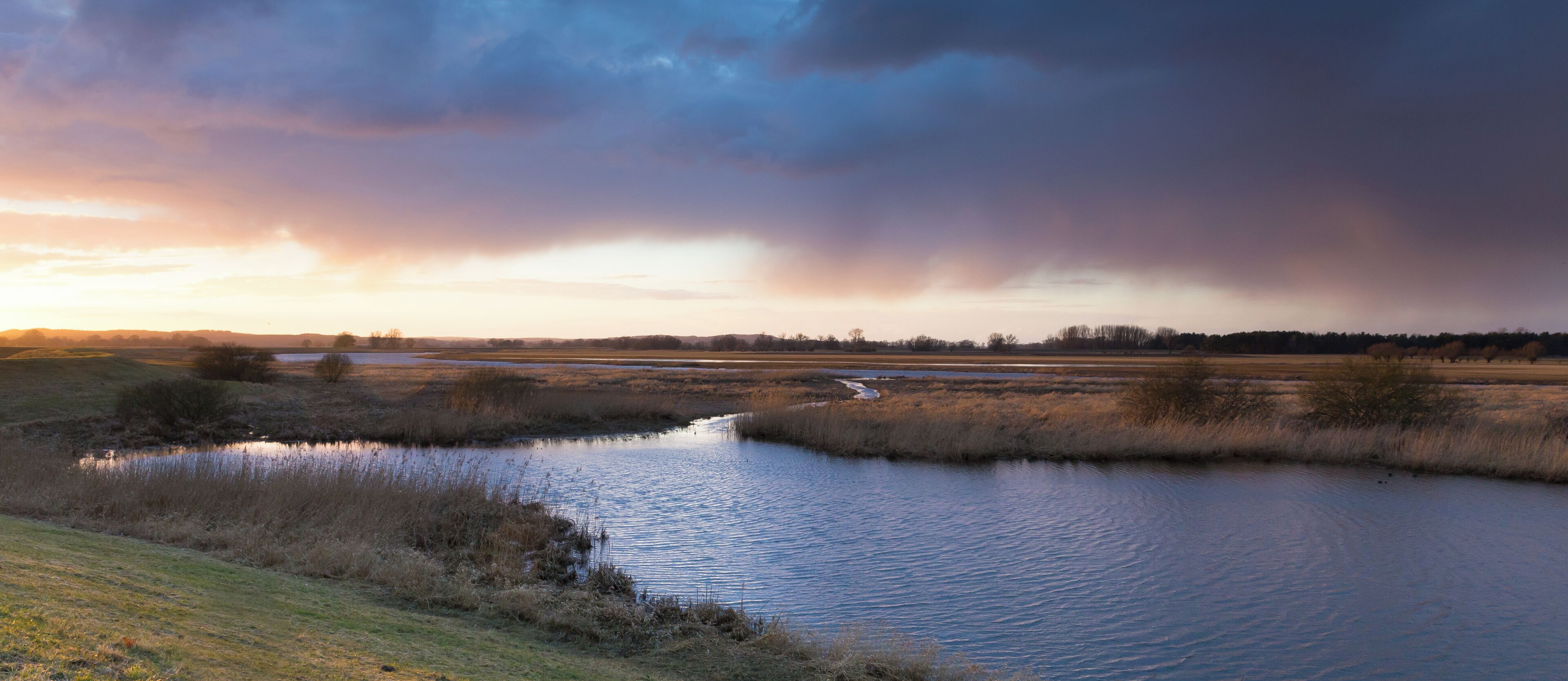 A river meandering through a flat landscape with sparse vegetation and a dramatic sky.