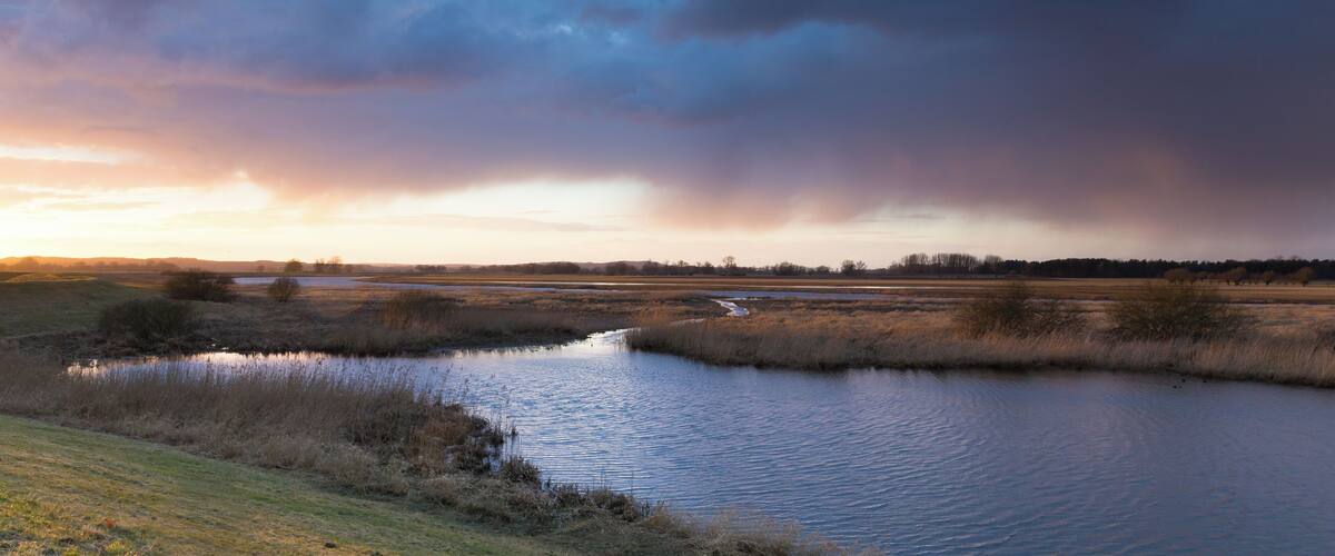 A river meandering through a flat landscape with sparse vegetation and a dramatic sky.