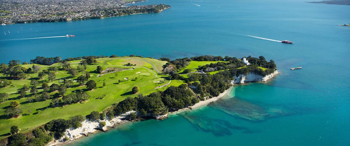 An aerial view of a coastal golf course with a clear blue sea, a sandy beach, and a distant city skyline.