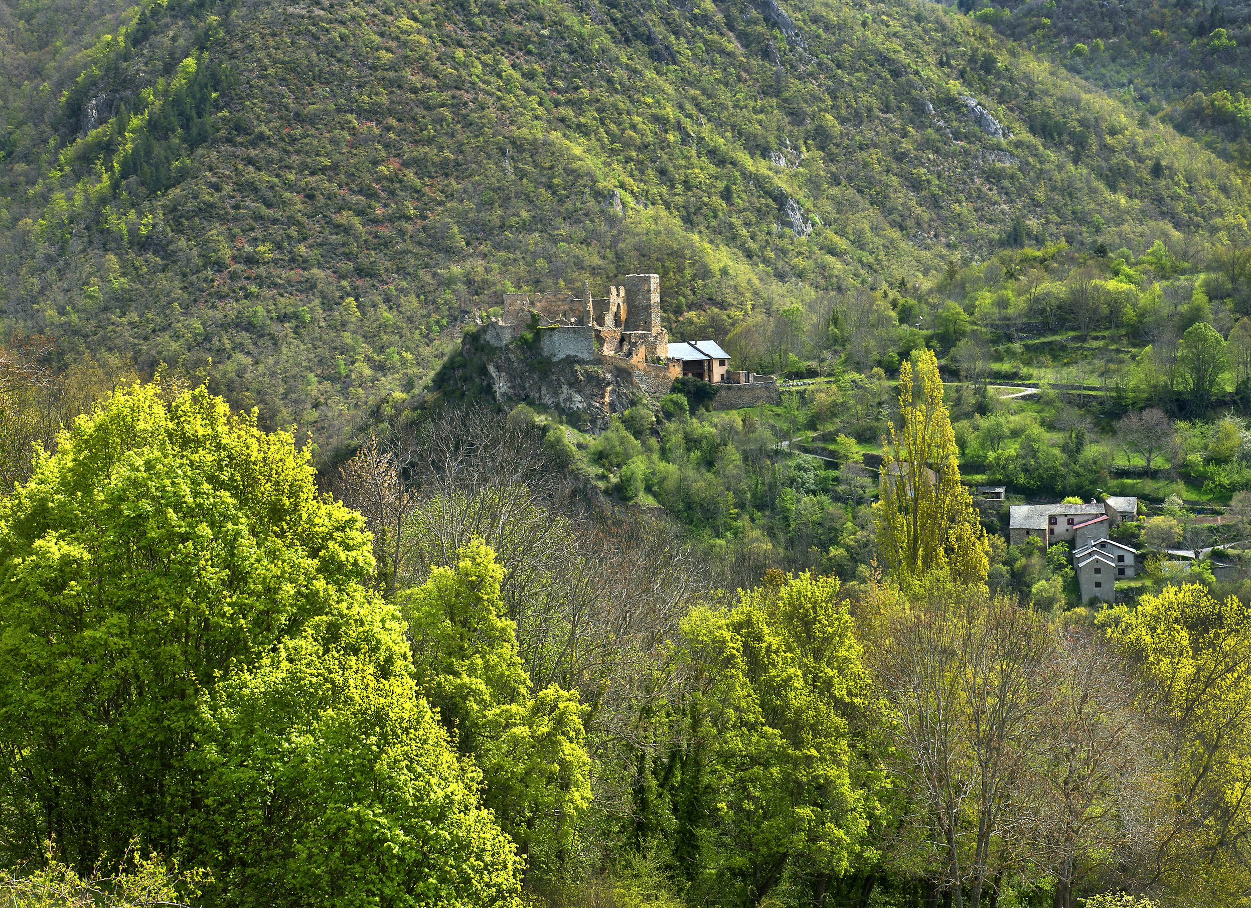 Un paysage montagneux avec une construction en pierre d’époque qui se dresse au sommet d’une colline, entourée d’une végétation luxuriante et surplombant un petit village.