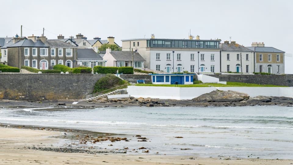 A coastal town with terraced houses, a rocky shoreline, and a small blue structure near the water.