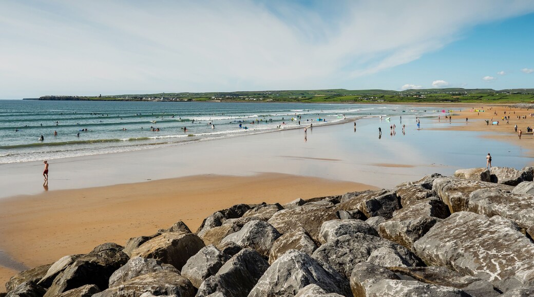 Yellow sand and blue ocean and sky. People swim in the background and surf on board. Lahinch beach in county Clare, Ireland. Warm sunny day. Irish landscape.