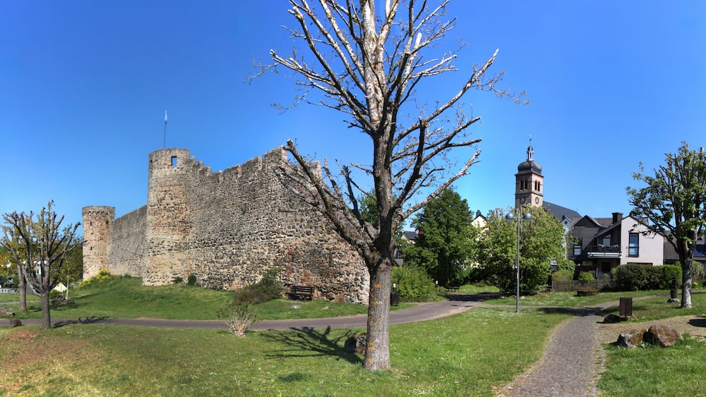A historic stone castle with a tower, surrounded by a park with pathways, benches, and modern houses.