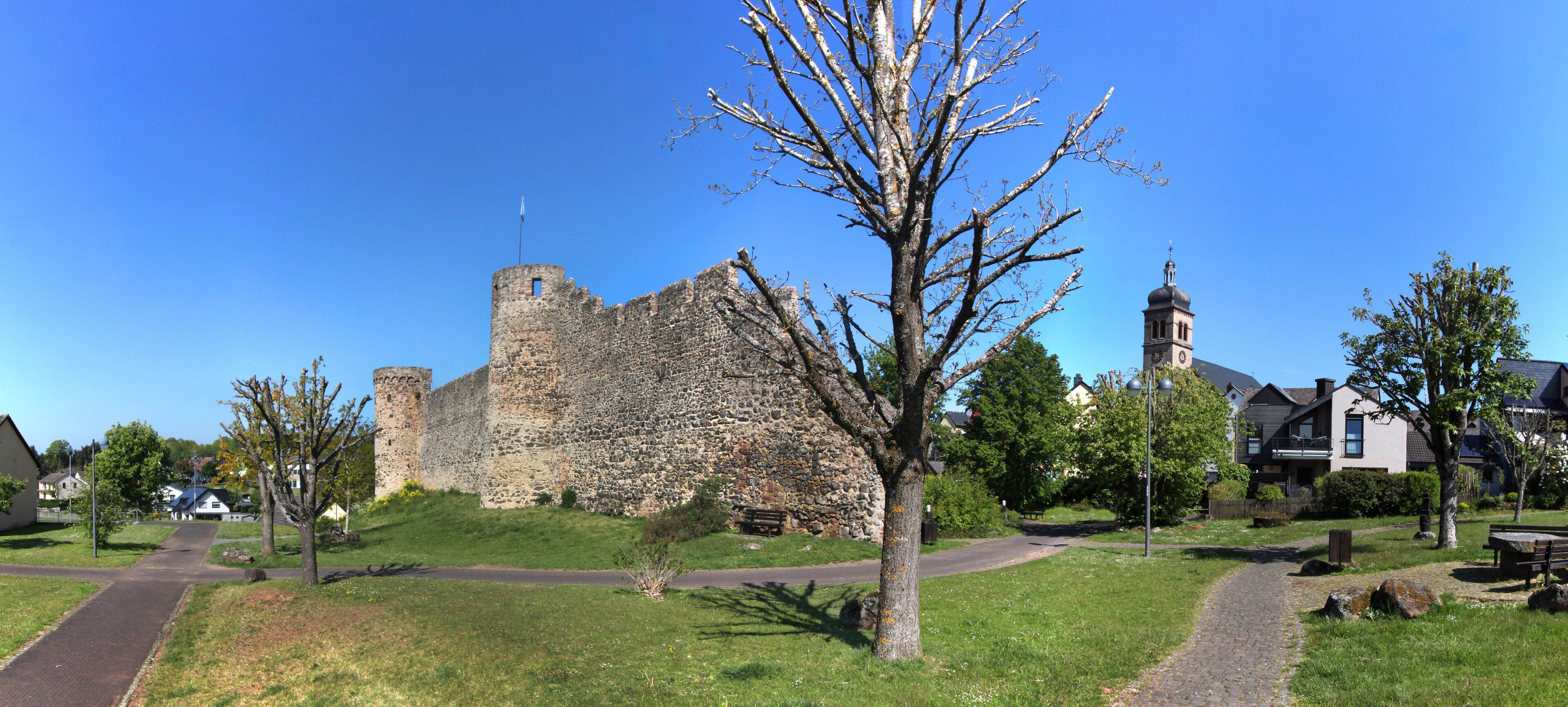 A historic stone castle with a tower, surrounded by a park with pathways, benches, and modern houses.
