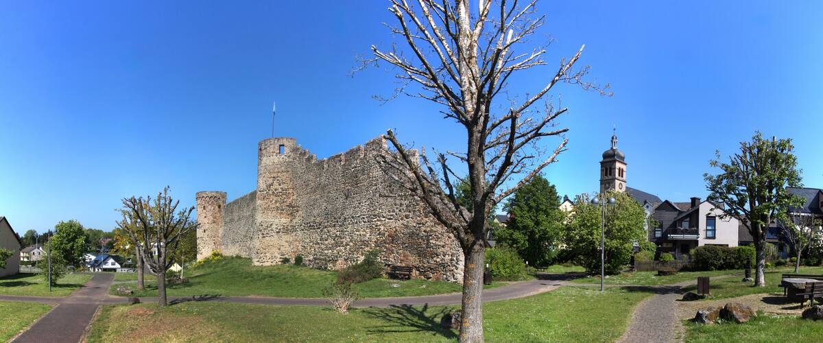 A historic stone castle with a tower, surrounded by a park with pathways, benches, and modern houses.
