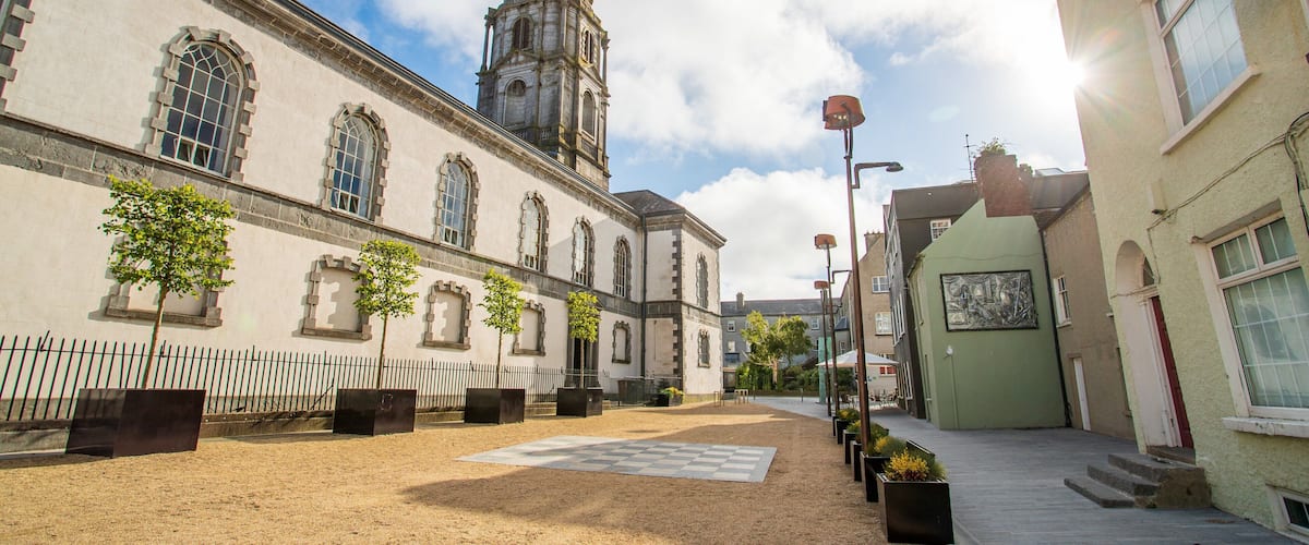 A historic building with arched windows and a tall spire, surrounded by a cobblestone square and modern buildings.