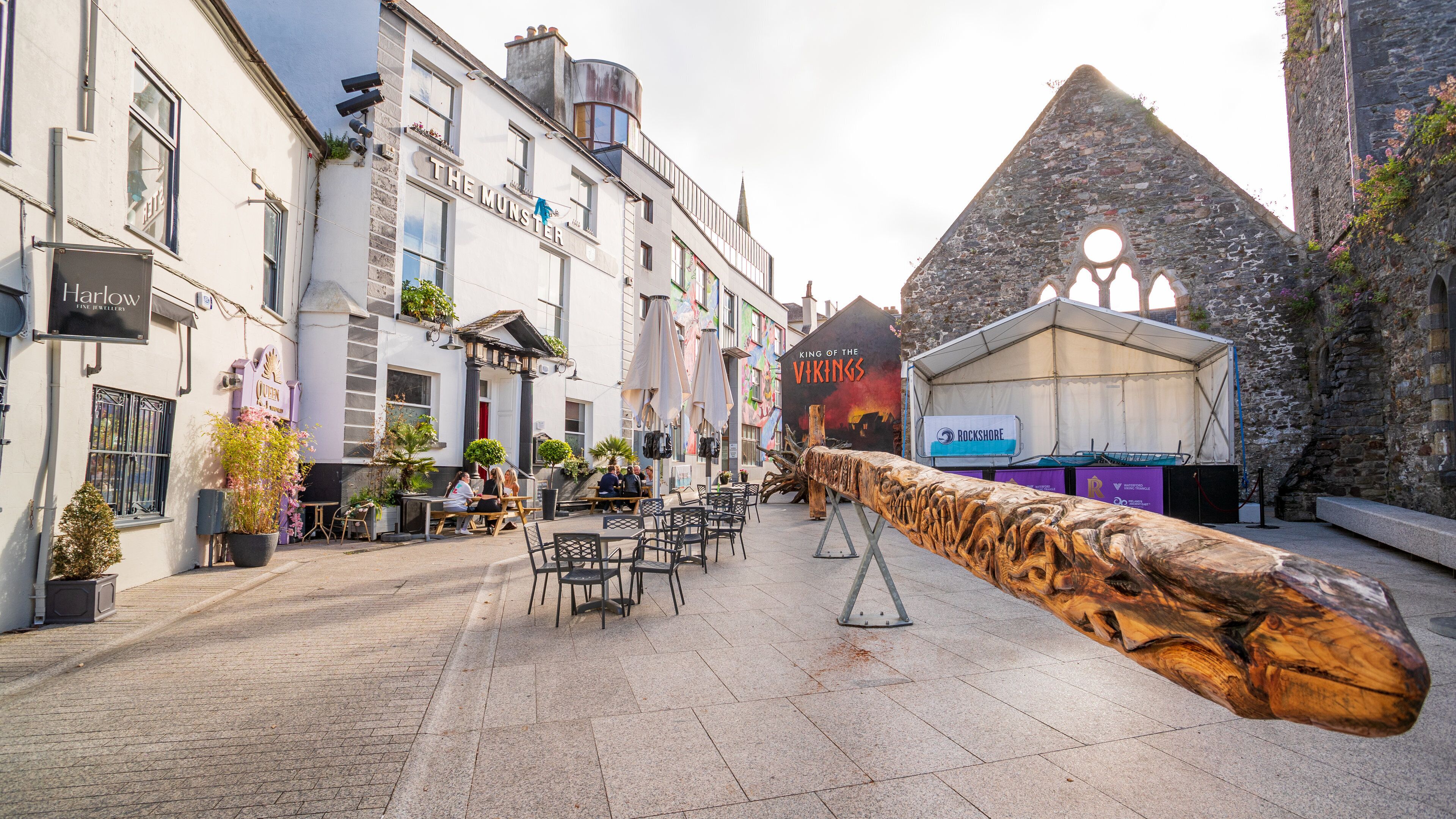 A cobblestone street with outdoor seating, a wooden log sculpture, and a building with a sign reading "The Munster."