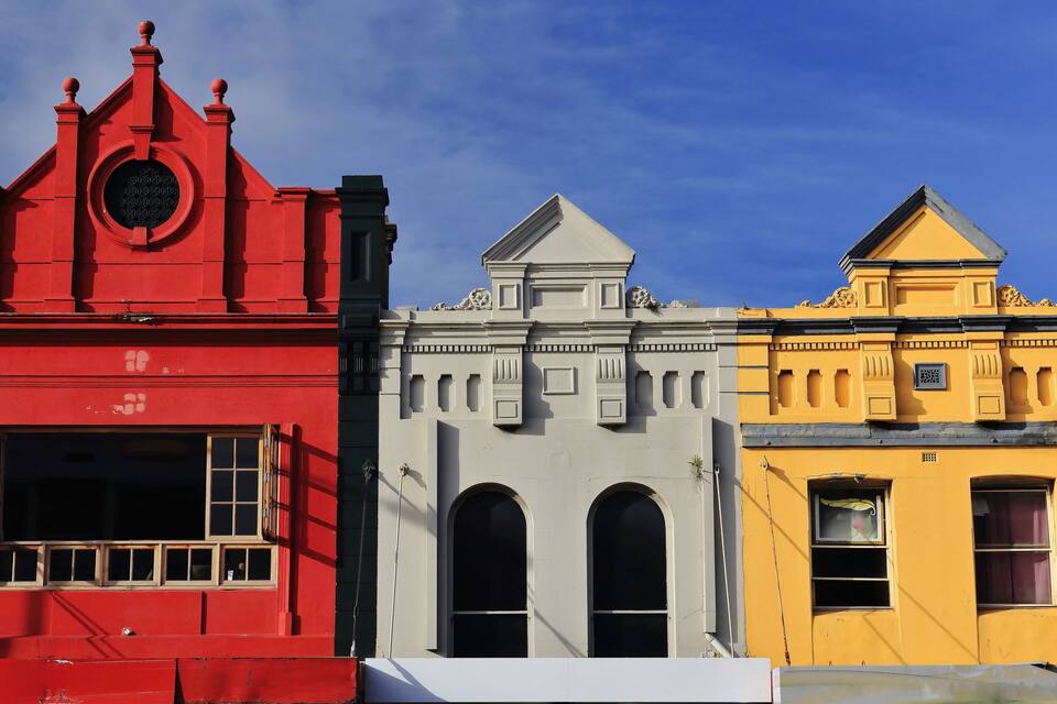 Una hilera de edificios coloridos con estilos arquitectónicos diferentes, incluyendo un edificio rojo con una ventana circular, uno gris con ventanas arqueadas y uno amarillo con detalles decorativos.