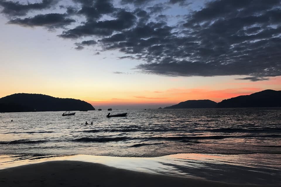 A sunset over a calm sea with boats and a sandy beach.