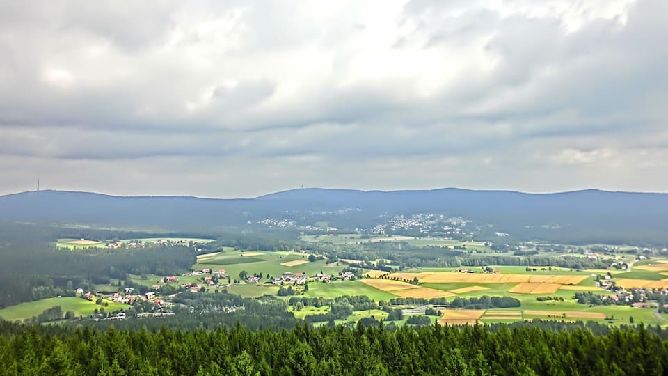 A rural landscape with rolling hills, scattered houses, and a dense forest in the foreground.